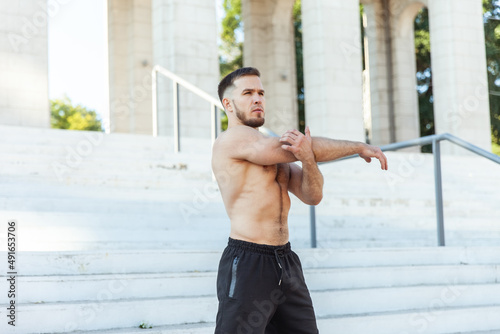 Young male athlete with a naked torso kneads the muscles doing stretching exercise while standing on the stairs outdoors