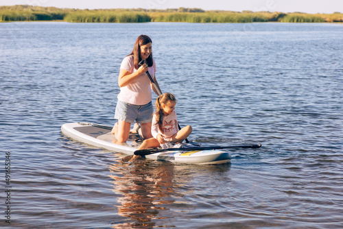 Middle-aged Caucasian mother with little daughter sup boarding both smiling and looking down at water. Active lifestyle and family time together. Teaching children to do sports. © Юля Бурмистрова