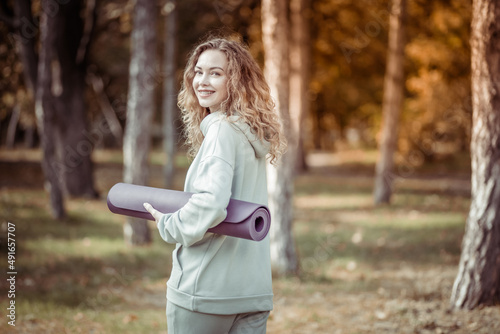 Cheerful smiling fit of a Caucasian woman with curly hair holding yoga mat in her hand in the park at bright sunny day. Healthy lifestyle. Outdoor workout