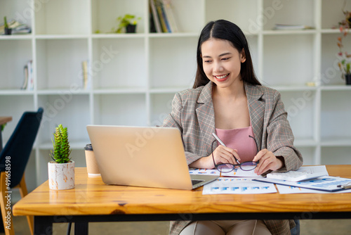 Portrait of smiling Asian businesswoman working at a trendy desk. Ecommerce Beautiful Asian businesswoman analyzing charts using laptop calculator at the office. © ArLawKa