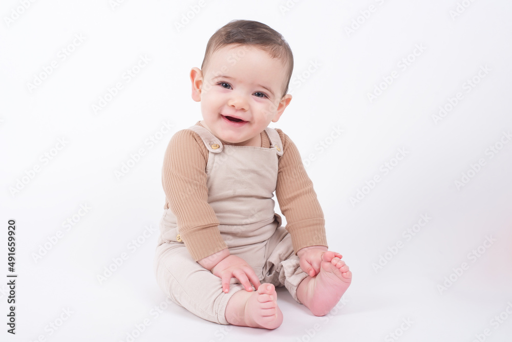 Adorable baby boy wearing beige overalls sitting on white background looking at camera and smiling. Adorable baby boy wearing beige overalls sitting on white background looking at camera and smiling.