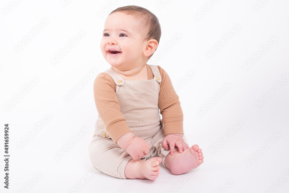Adorable seven month old baby boy wearing beige overalls sitting on the floor over white background looking aside and smiling. Adorable seven month old baby boy wearing beige overalls sitting on the floor over white background looking aside and smiling.