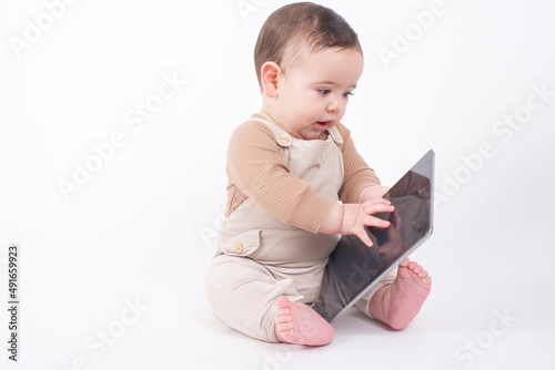 Adorable baby boy wearing beige overalls sitting on white background holding a tablet pad and watching streaming videos or cartoons. 