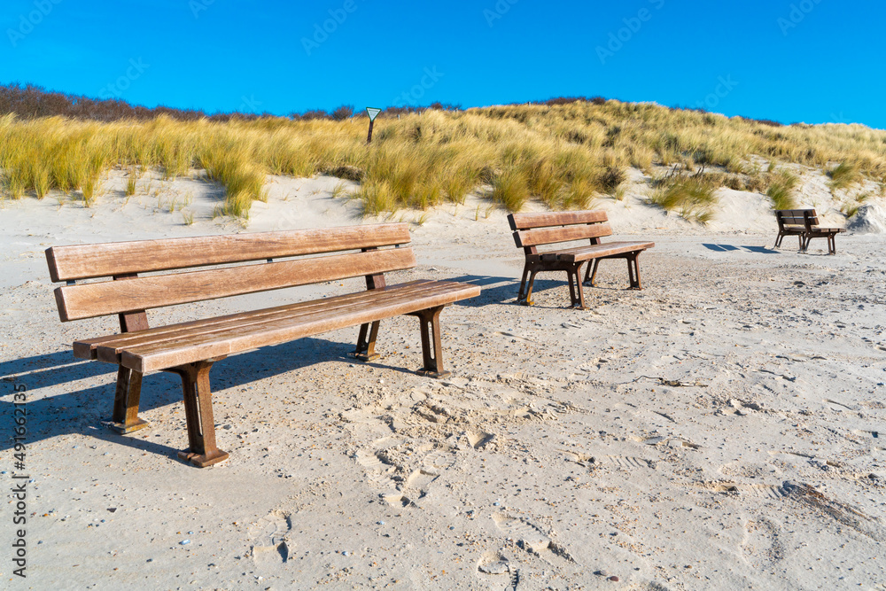 Three wooden benches on a sandy beach of the Dune island, Heligoland, Germany, on a beautiful sunny day of winter. Dunes with yellow grass in the back. Three wooden benches on a sandy beach of the Dune island, Heligoland, Germany, on a beautiful sunny day of winter. Dunes with yellow grass in the back.