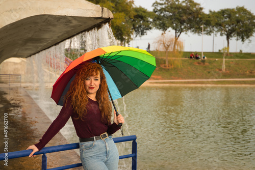 Young woman, red hair, freckles, with a rainbow umbrella, under a waterfall, in an outdoor park. Concept color, happiness, well-being, fun, rain.