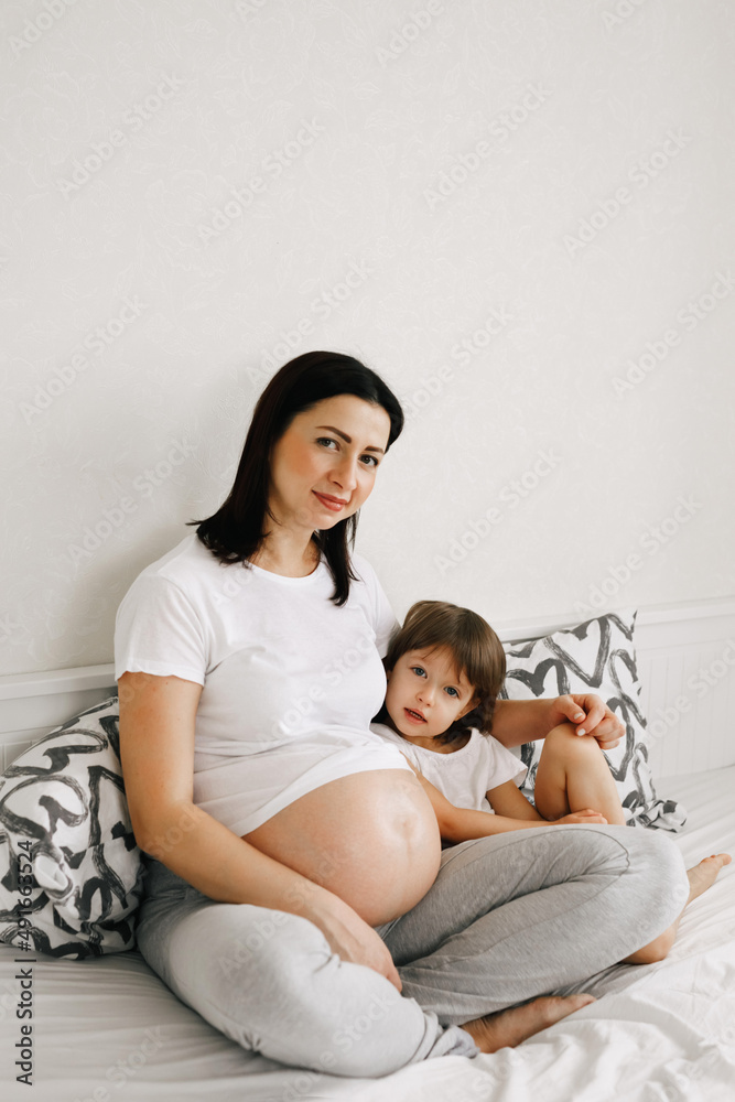 Pregnant expecting mother relaxing with little girl daughter in white bedroom interior at home. Waiting child, motherhood concept.