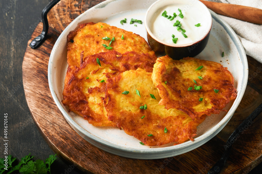 Potato pancakes. Fried homemade potato pancakes or latkes with cream and green onions in rustic plate on old wooden black table background. Rustic style. Healthy food. Top view. Potato pancakes. Fried homemade potato pancakes or latkes with cream and green onions in rustic plate on old wooden black table background. Rustic style. Healthy food. Top view.