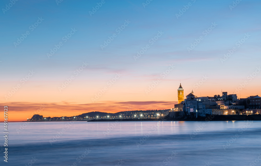 Fotografia de larga exposición de Sitges desde la playa con la iglesia iluminada al fondo en un atardecer, anochecer con cielo de colores azul, naranja y amarillo y unas ligeras nubes en el horizonte