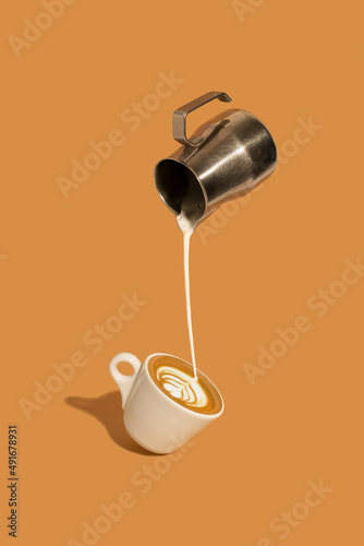 Pouring creamy milk from a stainless steel milk jug, and making late art espresso in a coffee cup on brown background.
