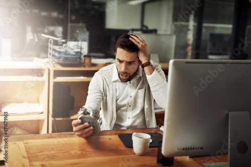 There just arent enough hours in the day. Shot of a handsome young businessman holding an alarm clock and looking stressed out while working late in an office.