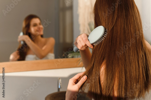Smiling pretty brunette in a towel combs her long shiny silky hair with a comb in the morning after cosmetic procedures, on the background of the bathroom.