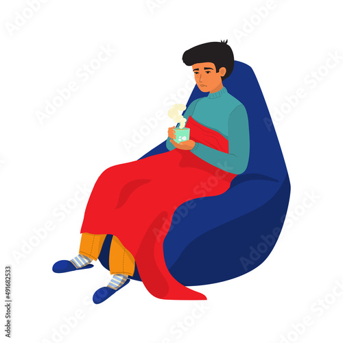 The man got sick and caught a cold. He sits on a bean bag wrapped in a blanket. drinking hot drink