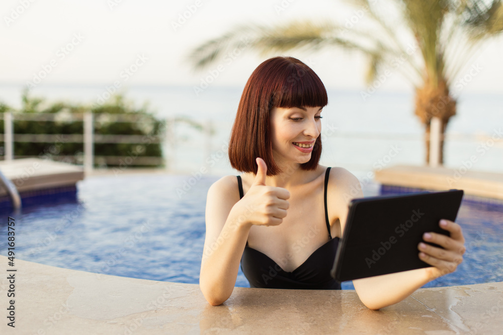Attractive joyful Caucasian woman wearing black swimsuit, using tablet pc and showing thumb up, while swimming at the resort pool. Summer and holiday concept.