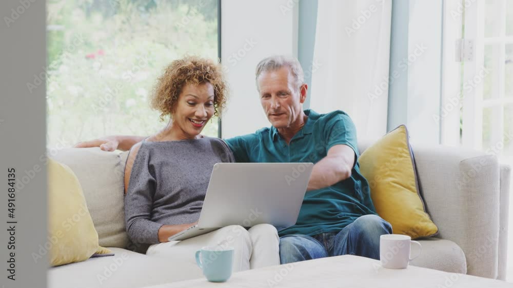 Smiling senior retired couple sitting on sofa at home booking holiday or shopping on laptop - shot in slow motion Smiling senior retired couple sitting on sofa at home booking holiday or shopping on laptop - shot in slow motion