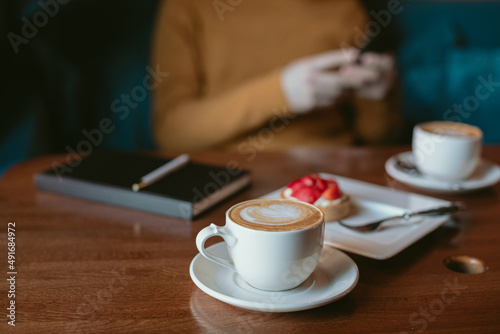 Cup of coffee, delicious cute strawberry tart, black notebook on wooden table, man checking his mobile phone on background. Man having a break in a coffee shop.