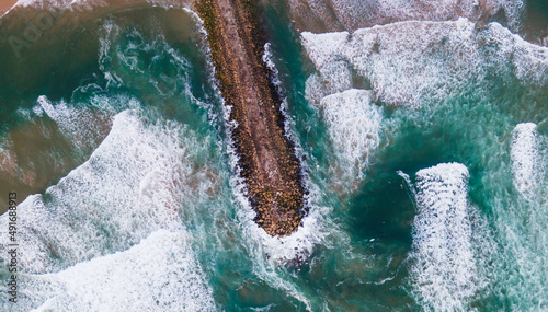 Aerial view of a majestic breakwater with rough rolling water waves rolling on the beach at Costa da Caparica, Setubal, Portugal.