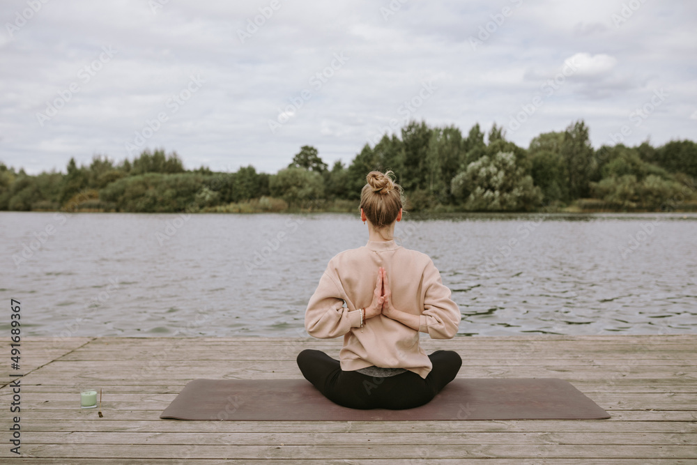 Young woman sitting on the exercise mat, meditating in the lotus pose during a yoga on the pier. Self-analysis and soul-searching concept. Zen hands. Young woman sitting on the exercise mat, meditating in the lotus pose during a yoga on the pier. Self-analysis and soul-searching concept. Zen hands.