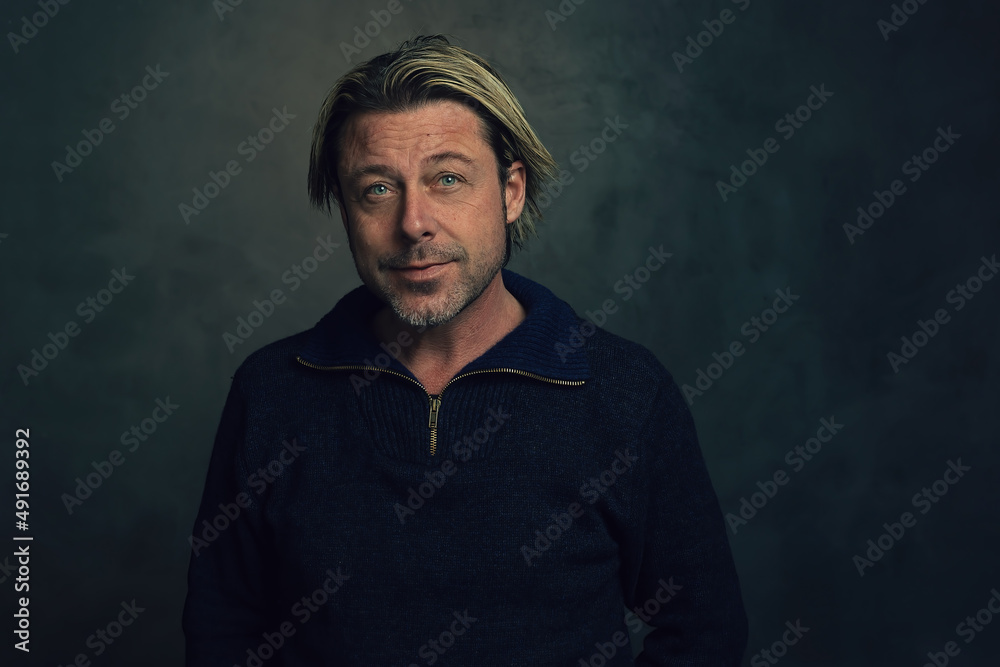 Rugged blond man with a stubble in a blue woolen sweater with zipper collar in front of a dark grey wall. Rugged blond man with a stubble in a blue woolen sweater with zipper collar in front of a dark grey wall.