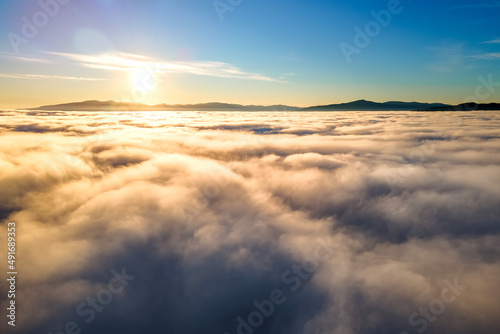 Aerial view of yellow sunset over white puffy clouds with distant mountains on horizon © bilanol
