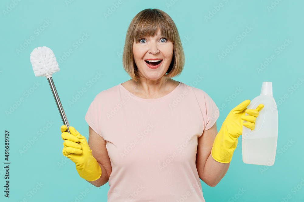 Elderly housewife woman 40s in pink t-shirt gloves hold toilet brush sponge detergent chemical bottle cleanser isolated on plain pastel light blue background. Housekeeping cleaning tidying up concept. Elderly housewife woman 40s in pink t-shirt gloves hold toilet brush sponge detergent chemical bottle cleanser isolated on plain pastel light blue background. Housekeeping cleaning tidying up concept.