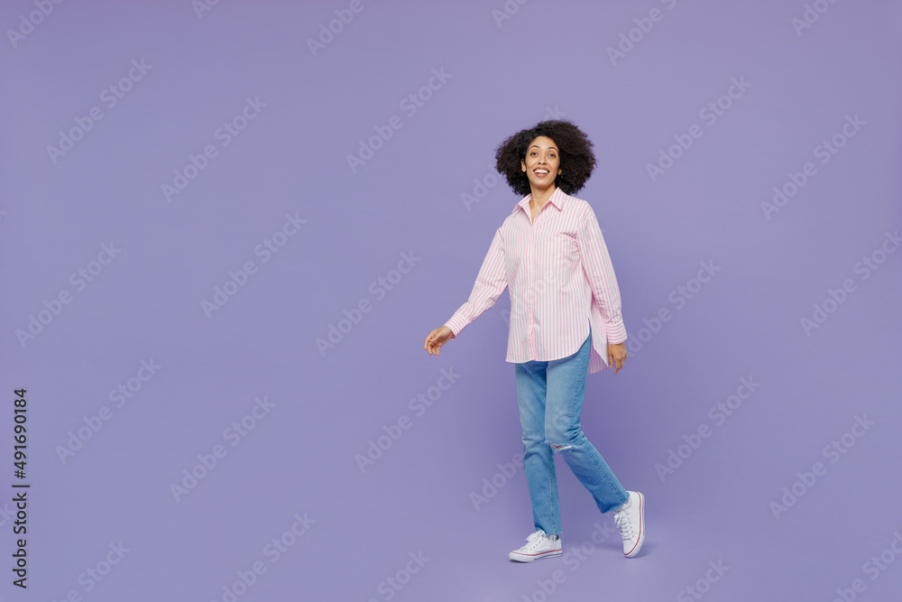 Full body smiling happy fun cheerful young woman of African American ethnicity 20s wear pink striped shirt walking going look camera isolated on plain pastel light purple background studio portrait. Full body smiling happy fun cheerful young woman of African American ethnicity 20s wear pink striped shirt walking going look camera isolated on plain pastel light purple background studio portrait.