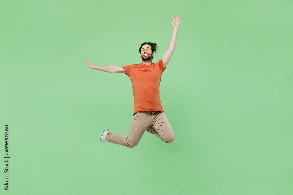 Full body young excited fun cool man 20s wear casual orange t-shirt jump high with outsretched hands isolated on plain pastel light green color background studio portrait. People lifestyle concept