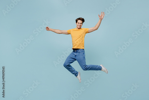 Full body overjoyed fun joyful cool young man 20s wearing yellow t-shirt jumping high with outstretched hands isolated on plain pastel light blue background studio portrait. People lifestyle concept.