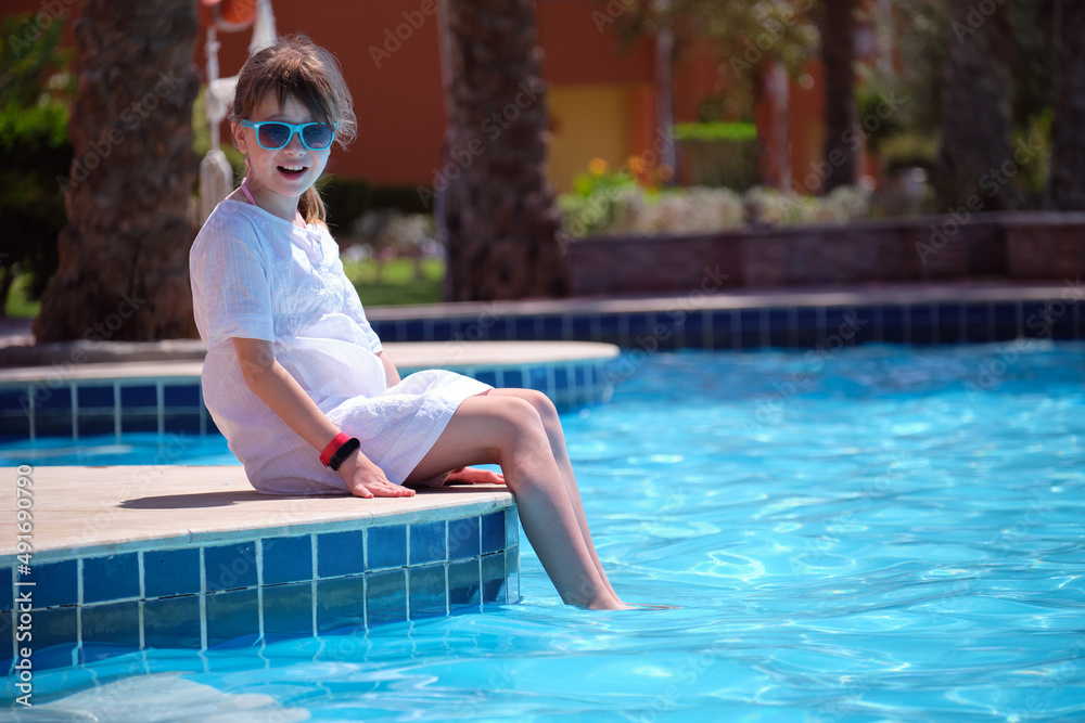 Portrait of happy child girl in white dress relaxing on swimming pool side on sunny summer day during tropical holidays