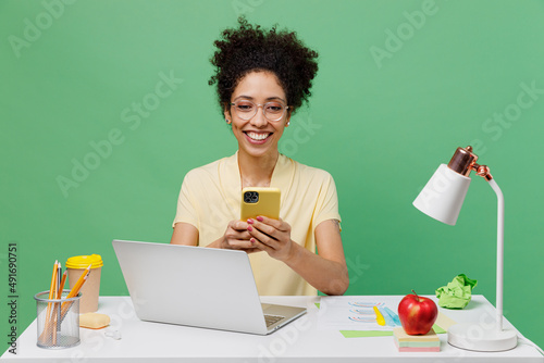 Young employee business woman of African American ethnicity in shirt sit work at white office desk with pc laptop use mobile cell phone isolated on plain green background. Achievement career concept.