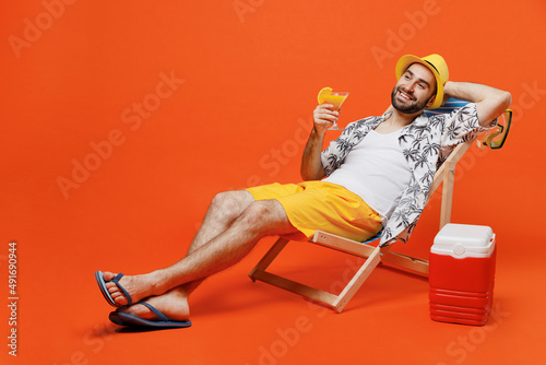Canvas Print Young happy cool tourist man 20s wear beach shirt hat lie on deckchair near fridge drink exotic cocktail isolated on plain orange background studio portrait