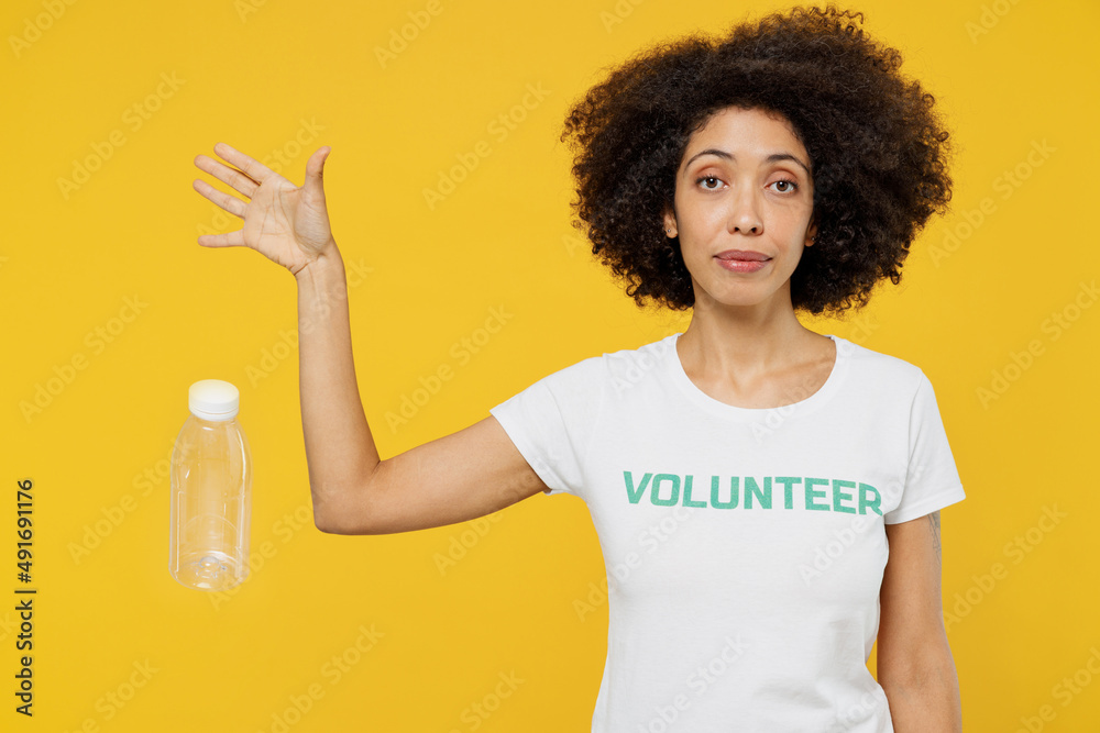 Young sad woman of African American ethnicity in white volunteer t-shirt throw out refuse plastic pet bottles isolated on plain yellow background. Voluntary free work assistance help charity concept. Young sad woman of African American ethnicity in white volunteer t-shirt throw out refuse plastic pet bottles isolated on plain yellow background. Voluntary free work assistance help charity concept.