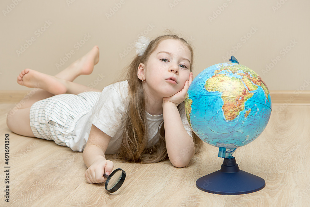 pensive little girl lies on the floor next to a globe, holds a magnifying glass in one hand, put the other under her head and argues. The concept of a smart child, education, plans for the future pensive little girl lies on the floor next to a globe, holds a magnifying glass in one hand, put the other under her head and argues. The concept of a smart child, education, plans for the future