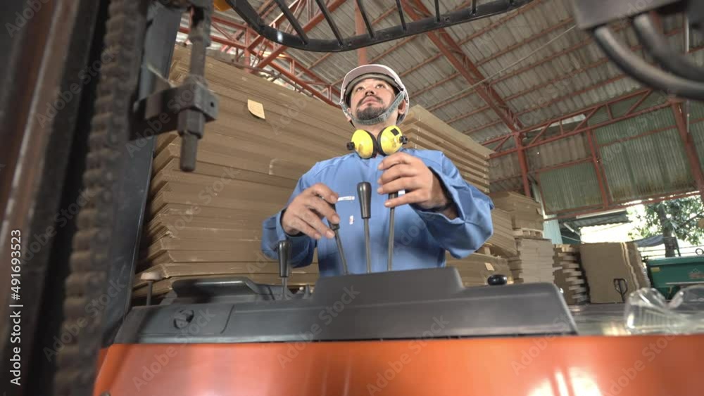 Low angle, front view of Asian male factory worker with beard in a helmet and ear muffs around his neck sitting controlling lifting levers on a forklift truck while looking forward in a paper factory.