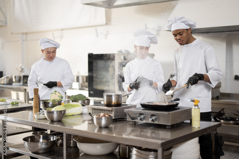 Multiracial team of professional cooks in uniform preparing meals for a restaurant in the kitchen. Latin guy frying meat, european cooks making sauce and asian chef managing the process. Teamwork and Multiracial team of professional cooks in uniform preparing meals for a restaurant in the kitchen. Latin guy frying meat, european cooks making sauce and asian chef managing the process. Teamwork and