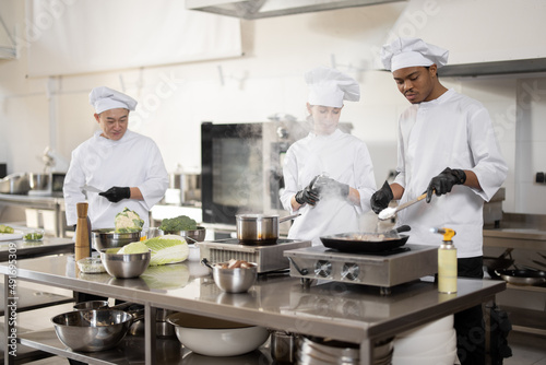 Multiracial team of professional cooks in uniform preparing meals for a restaurant in the kitchen. Latin guy frying meat  european cooks making sauce and asian chef managing the process. Teamwork and