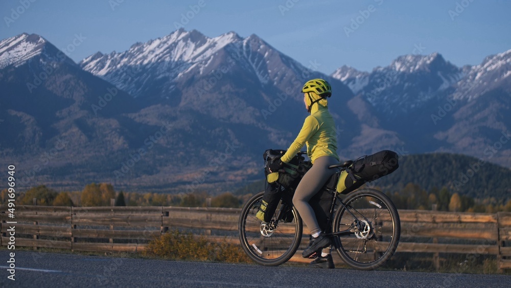 The woman travel on mixed terrain cycle touring with bikepacking. The traveler journey with bicycle bags. Sport tourism bikepacking, bike, sportswear in green black colors. Mountain snow capped.