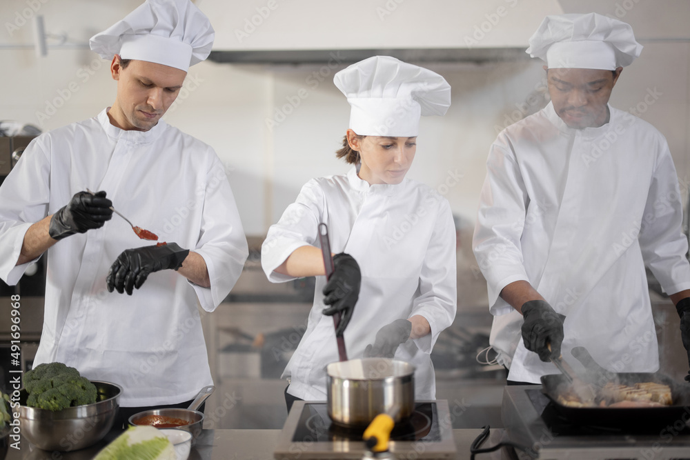 Multiracial team of three cooks in uniform cooking together in the professional kitchen. Latin cook frying meat and european guys making sauce. Concept of teamwork at restaurant Multiracial team of three cooks in uniform cooking together in the professional kitchen. Latin cook frying meat and european guys making sauce. Concept of teamwork at restaurant