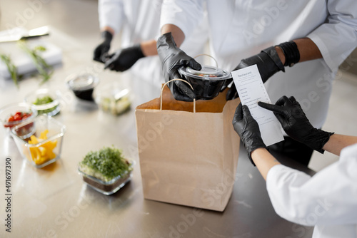 Cooks in protective gloves prepare take away food and packing them into paper bag for delivery, close-up on food. Concept of cooking food at dark kitchen for delivery photo