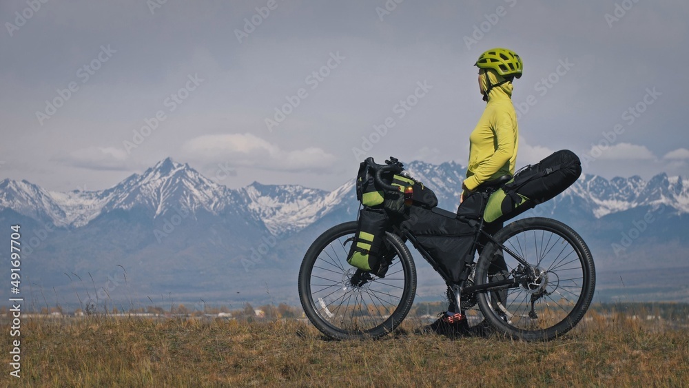 The woman travel on mixed terrain cycle touring with bikepacking. The traveler journey with bicycle bags. Sport tourism bikepacking, bike, sportswear in green black colors. Mountain snow capped. The woman travel on mixed terrain cycle touring with bikepacking. The traveler journey with bicycle bags. Sport tourism bikepacking, bike, sportswear in green black colors. Mountain snow capped.