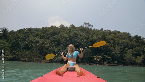 Attractive sportive blonde woman with sunglasses rows pink plastic kayak putting up paddle on sea against hills with wild jungles, blue sky. Traveling to tropical countries. Girl is sailing on kayak. © ivandanru
