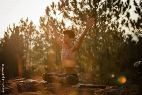 Young man doing yoga at the sunrise on a top of a mountain next the trees over the rocks with arm wide open