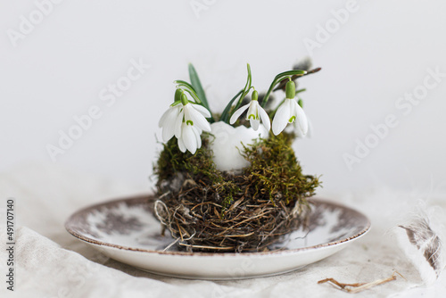 Stylish Easter table setting. Natural egg with snowdrops in nest on vintage plate and napkin on white wood. Modern Easter table rustic decoration