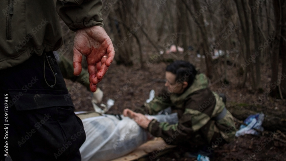 Hand close-up with blood on the fingers and a ring. Blurred figures of people in the forest. First aid in an extreme situation. Saving a life in a military conflict. Tactical medic, victim, rescue.