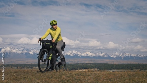 The man travel on mixed terrain cycle touring with bikepacking. The traveler journey with bicycle bags. Sport tourism bikepacking, bike, sportswear in green black colors. Mountain snow capped.