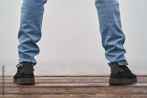 Into the unknown. Detail of legs in jeans and sneakers in front of ocean with fog over wood
