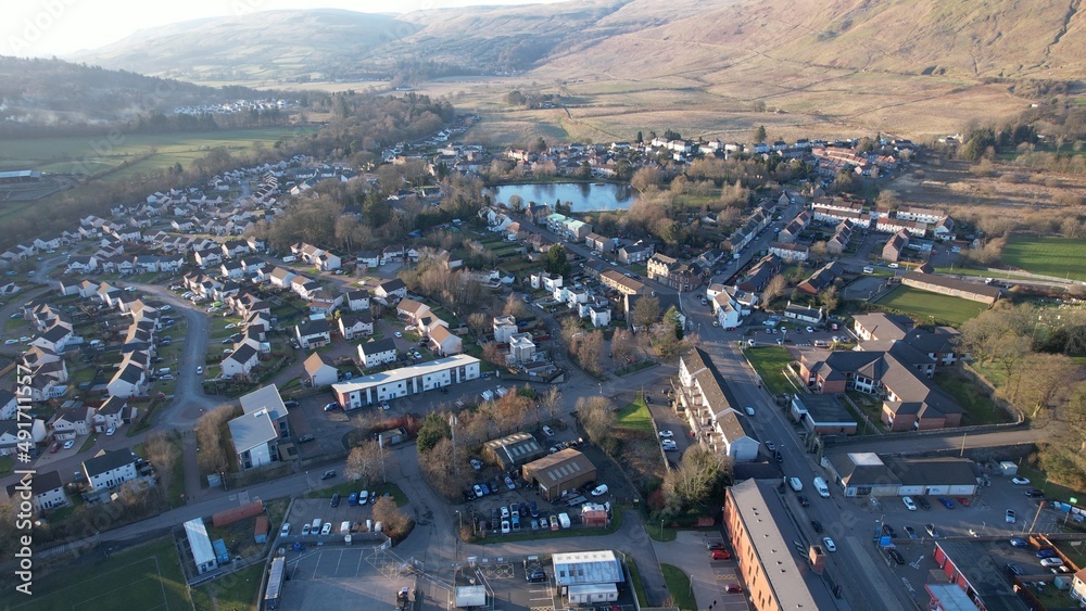 Fototapeta premium Low level aerial image of a Scottish village and glen surrounded by hills. A bright sunny day with a low sun casting long shadows. Village pond in the middle distance surrounded by houses.