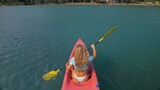 Seductive blonde woman in blue swimsuit sails on pink plastic kayak along azure sea at exotic resort backside upper above view. Traveling to tropical countries. Girl is sailing on kayak in ocean.