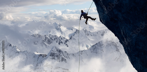 Adult adventurous man rappelling down a rocky cliff. Extreme adventure composite. 3d rendering mountain artwork. Aerial background landscape from British Columbia, Canada. Cloudy Sky