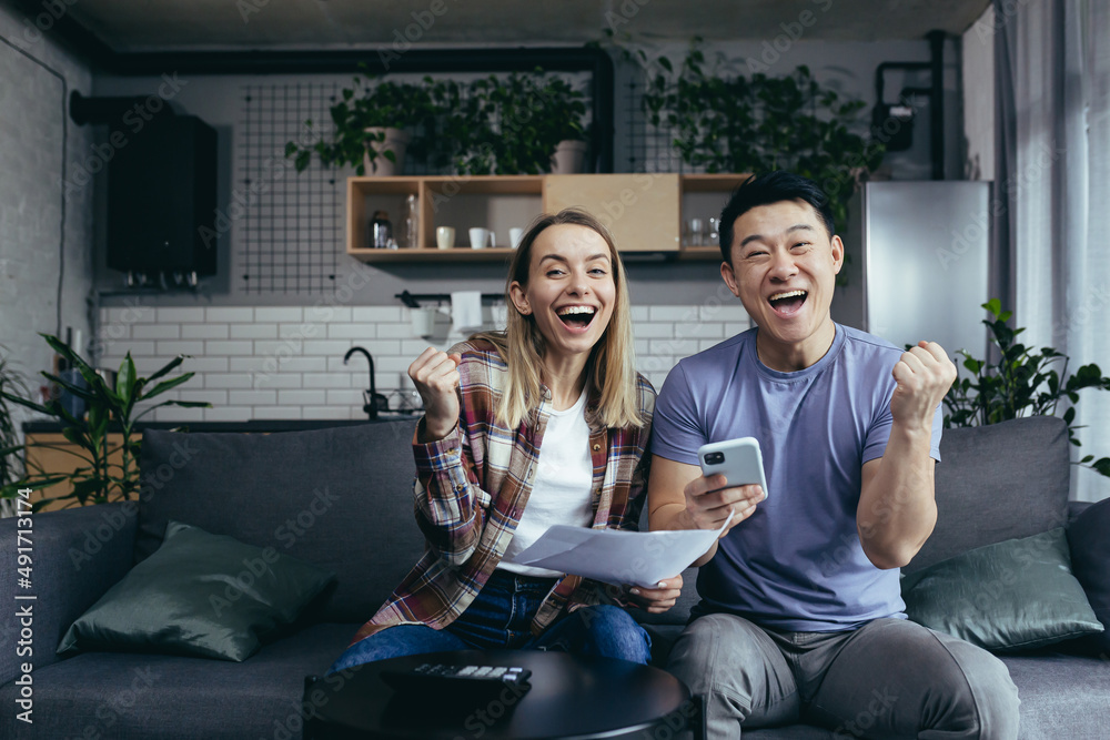 homework paper, multiracial couple, man and woman, pay bills by phone and app, sit at home on the couch, happy and smiling