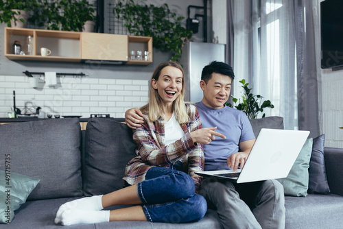 Young couple Asian man and woman at home sitting on sofa together using laptop, happy and smiling multiracial family looking at camera